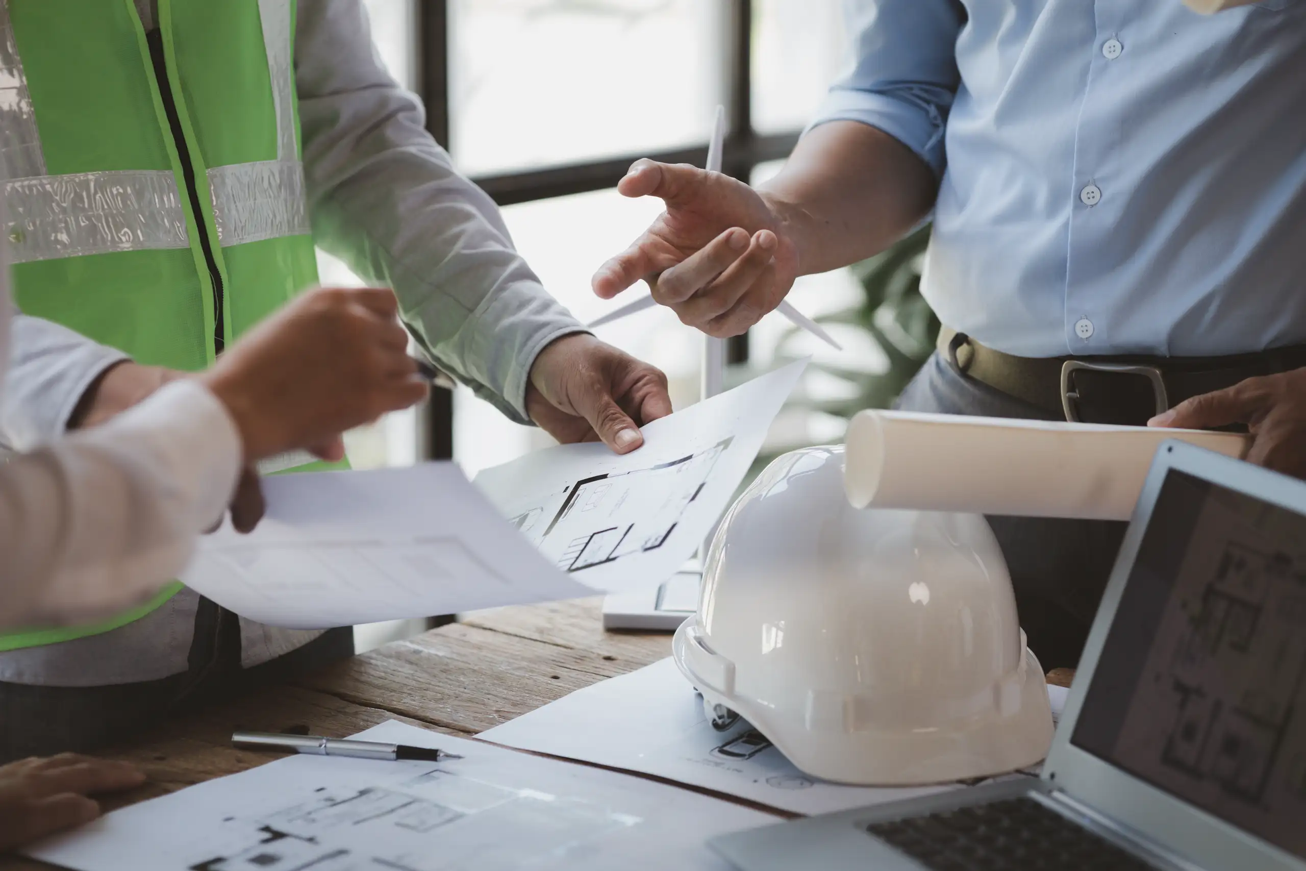 Mitarbeiter in Bauleitung besprechen Architekturpläne mit Schutzhelm auf Holztisch