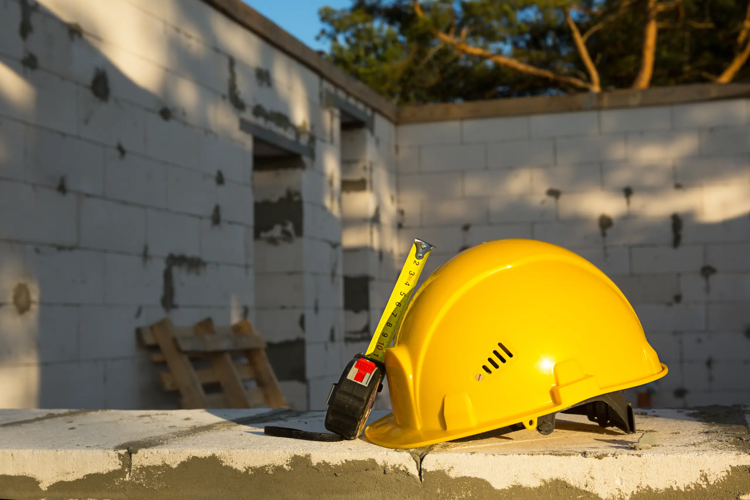 Gelber Schutzhelm mit ausziehbarem Maßband auf roher Betonplatte vor eingeschossigem Mauerwerk im Rohbau