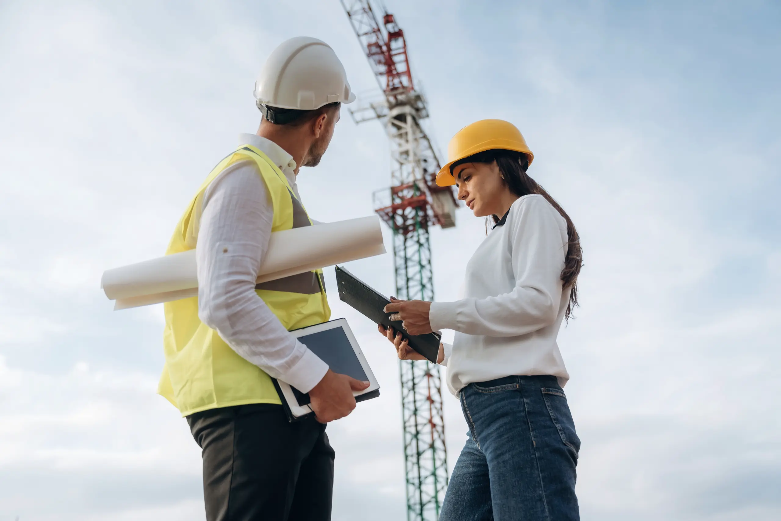 Bauleitung vor Ort: Ein Bauleiter und eine Projektmanagerin prüfen Pläne vor einem Kran auf der Baustelle