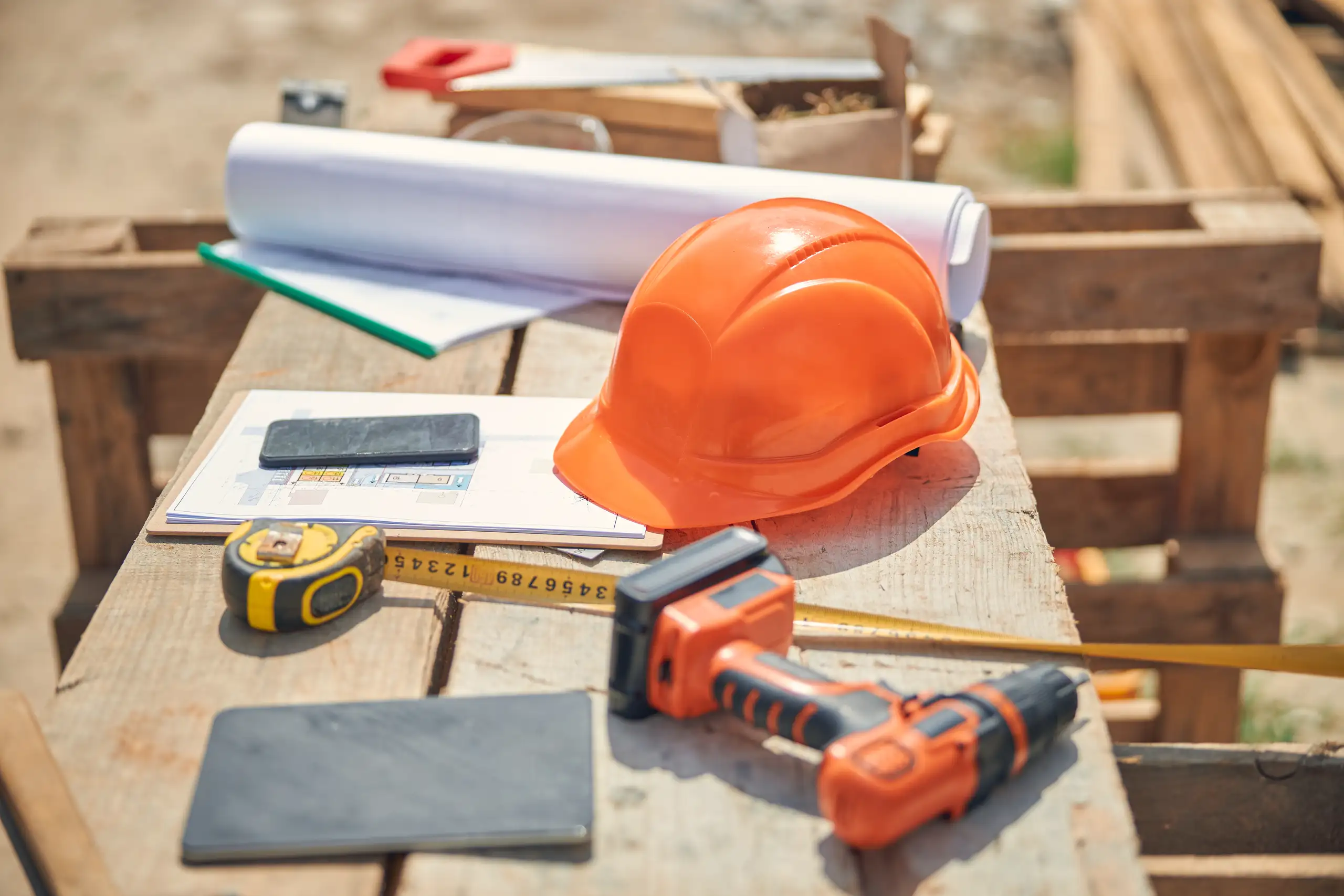 Auf dem Rohbau-Tisch liegen Baupläne, Bauhelm, Maßband und Akku-Bohrschrauber als Symbol für Bauleitung